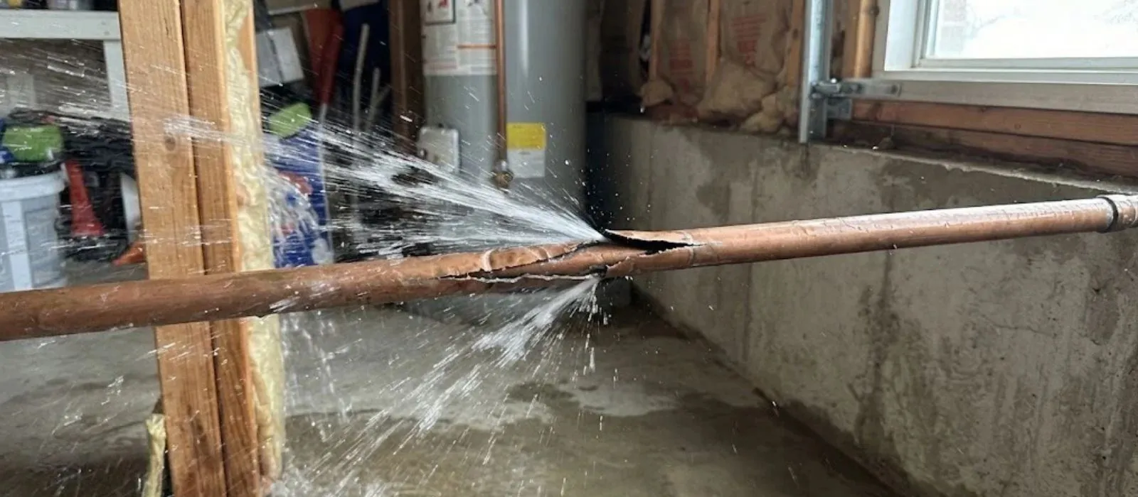 Burst copper pipe spraying water across a basement beside a water heater